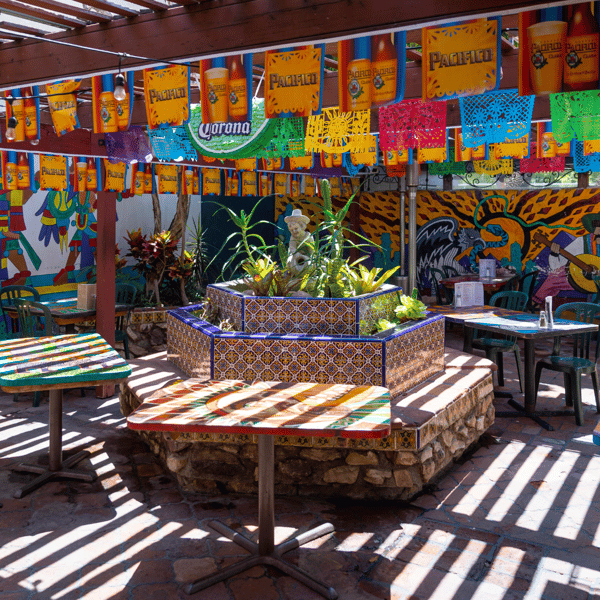A colorful outdoor dining area with vibrant paper banners, a tiled water fountain, and tables under striped sunlight.