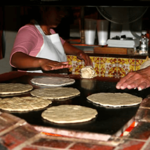 A person cooks tortillas on a griddle, while another prepares dough in the background. The scene is warm and rustic.