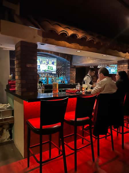 A cozy bar setting with dim lighting, showing a bartender serving drinks to two patrons sitting at the bar, with a TV screen in the background.