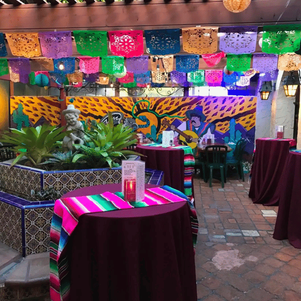 Colorful Mexican patio with papel picado banners, tiled fountain, mural, and tables draped in serapes with candles.