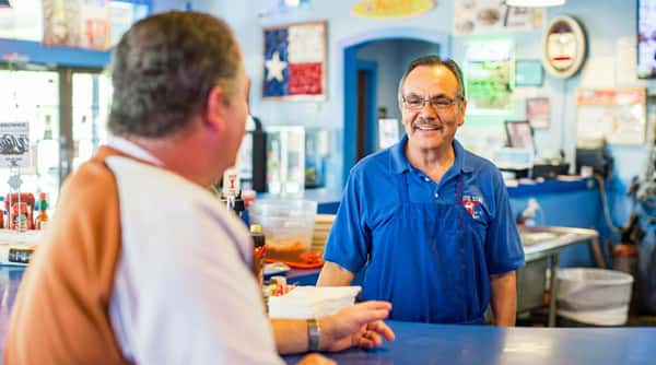 Man in blue apron smiling at another man across a blue counter in a restaurant