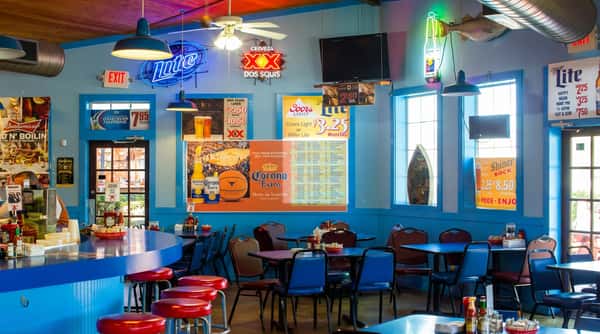 Interior of a bar or restaurant with blue walls, red stools at a counter, tables with blue chairs, and sports-themed beer advertisements.