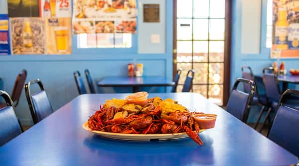 Five men in aprons stand behind a blue table with a plate of crawfish, corn, and potatoes.