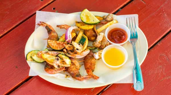 Bacon-wrapped shrimp skewers with seasoned zucchini, yellow squash, mushrooms, and onion, served with dipping sauces and a fork on a red table.