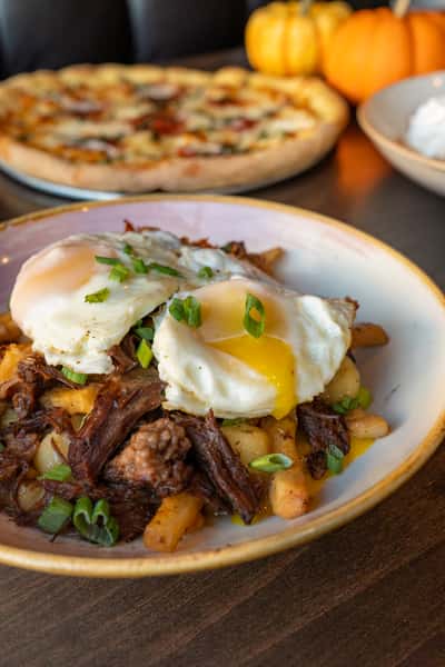 Welly's short rib hash and crispy potatoes topped with two runny fried eggs, with a pizza and fall pumpkins blurred in the background.