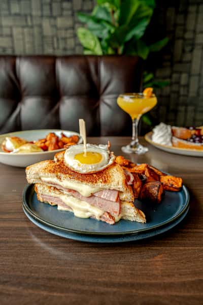 Grilled Croque Madame sandwich topped with a fried egg, served with roasted potatoes and a bright yellow brunch cocktail in a dark restaurant setting.