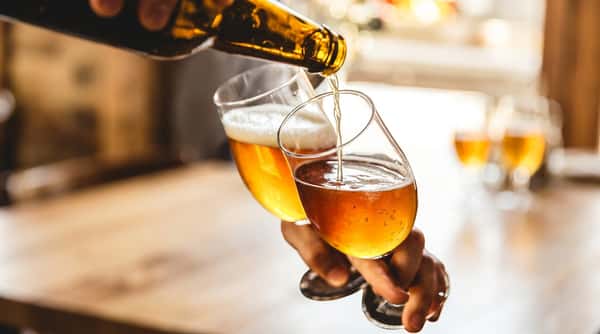 A person pours golden beer from a brown bottle into a tilted glass, forming foam.