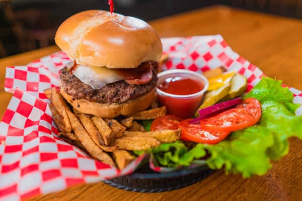 A large cheeseburger served on a wooden board with a side of waffle fries. The burger features a thick beef patty topped with melted cheese inside a shiny toasted bun held together by a skewer. A small cup of ketchup sits beside the fries, along with fresh lettuce, sliced tomatoes, and pickles for garnish. The red checkered paper liner and warm restaurant lighting create a classic American pub-style presentation.