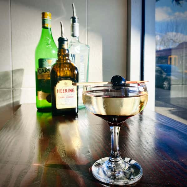 Image Description: Our cocktail, the Bettie Paige, garnished with a cherry. There are three bottles of liquor in the background.