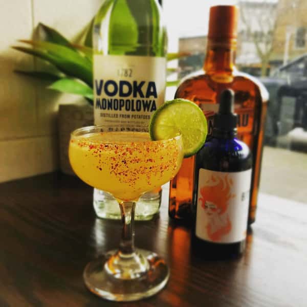 Image Description: An orange cocktail garnished with a tajin rim and a lime wheel is in the front, with three different liquor bottles and a fake plant in the background.