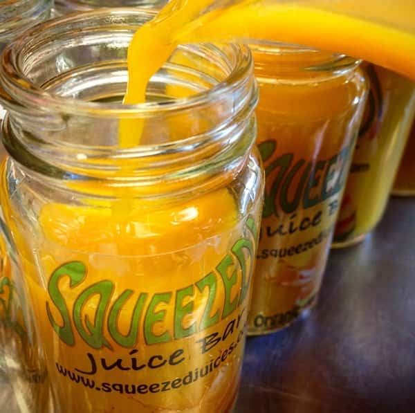 Golden orange juice being poured into several glass jars labeled "Squeezed Juice Bar" with green text.