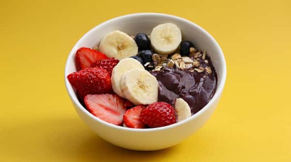 Acai bowl topped with banana slices, strawberries, granola, almonds, and honey drizzle in a clear container on a bold yellow background