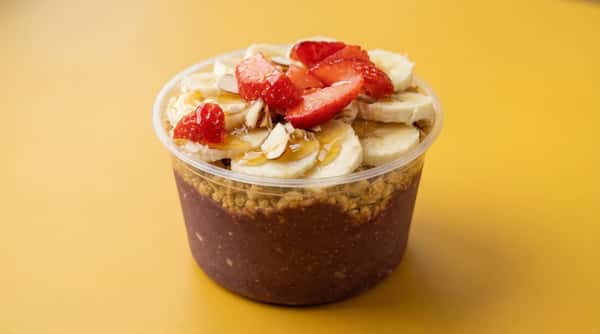 Acai bowl topped with fresh strawberries, banana slices, granola, almonds, and honey drizzle on a vibrant yellow backdrop