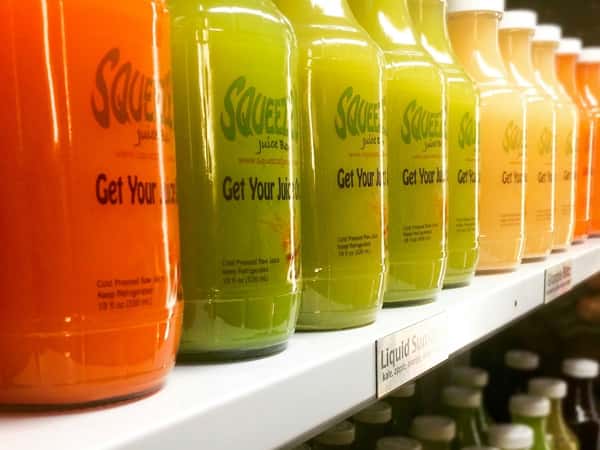 Row of colorful cold-pressed juices in glass bottles on display shelf, featuring green, citrus, and orange blends at a modern juice bar