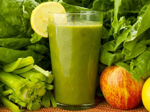 Green smoothie in a glass with a lemon slice, surrounded by lettuce, celery, an apple, and a lemon.