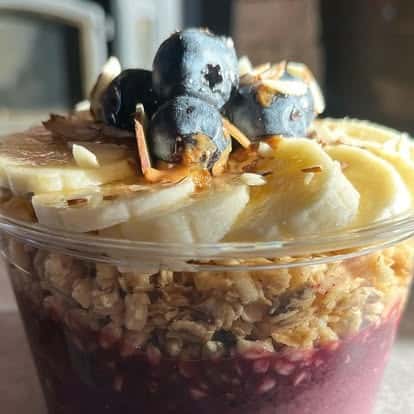 Acai bowl with granola, sliced bananas, blueberries, and strawberries in a clear plastic cup.