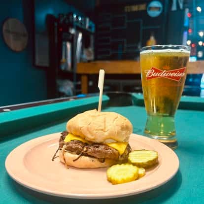 Cheeseburger with melted cheddar and pickle slices served with a cold draft beer beside a pool table in a neighborhood pub
