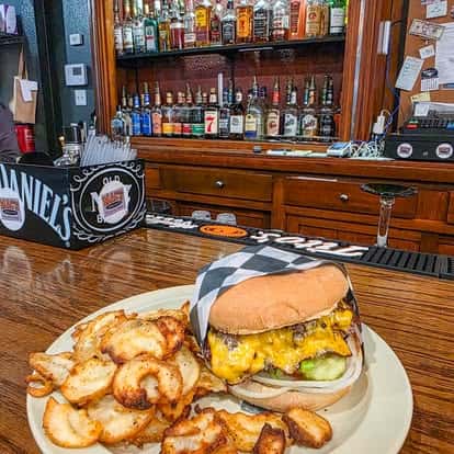 A juicy burger topped with cheese served on a white plate alongside a side of crispy seasoned potato wedges, with a bar interior visible in the background