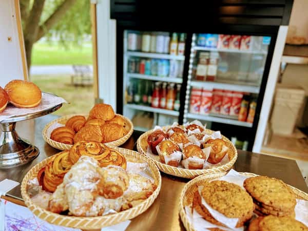 A display of baked goods, including baskets of croissants and cookies, arranged on a table near a glass-door beverage fridge.
