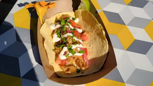 A soft taco filled with meat and topped with white sauce and cilantro, served in a cardboard boat on a geometric patterned table.