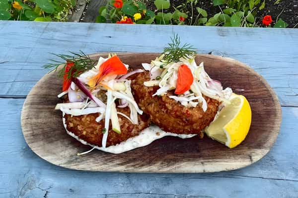 Two golden-fried fish or crab cakes topped with coleslaw and a tomato slice, served on a rustic wooden board.