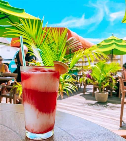 A layered red and white frozen cocktail with an orange slice and straw on a sunny tropical patio with green umbrellas and palm trees.