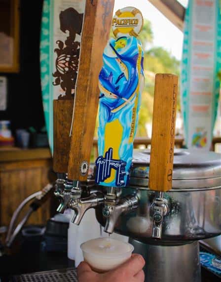 A hand fills a plastic cup with foamy draft beer from a vibrant Guy Harvey Pacifico tap handle at a beach-themed bar.