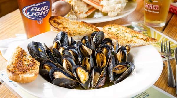 Bowl of steamed mussels with garlic bread, a glass of Bud Light, and a crab dish in the background.