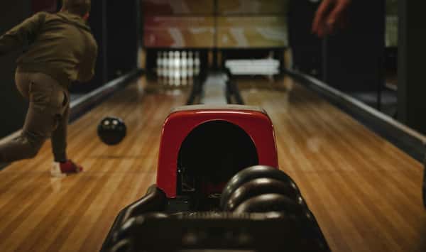 Person bowling on a wooden lane, ball rolling toward pins with a red ball return in the foreground.