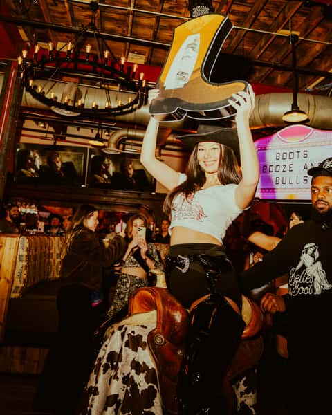 Guest riding a mechanical bull while holding an oversized cowboy boot prop inside a lively Western-themed bar with country nightlife energy.