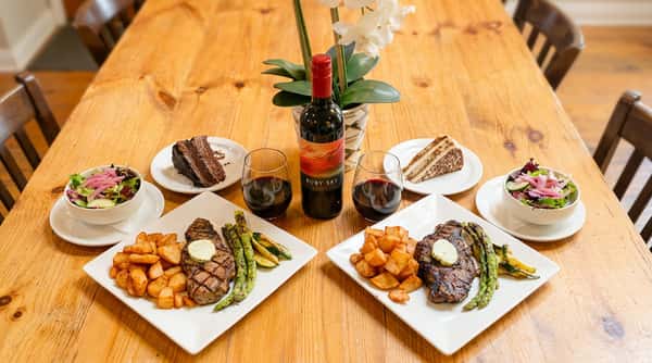 Two steak dinners with potatoes, asparagus, zucchini, salads, red wine, and slices of chocolate cake and tiramisu on a wood table.