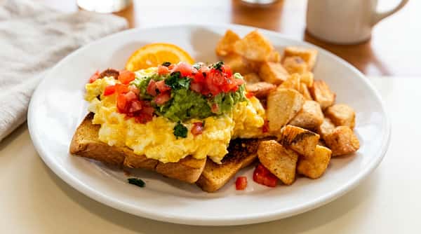 Scrambled eggs on toast with guacamole, diced tomatoes, a side of breakfast potatoes, and an orange slice.