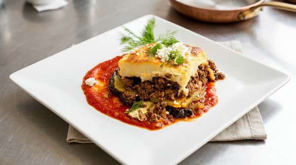 Moussaka with eggplant, minced meat, and béchamel sauce, garnished with feta and dill on a white plate.