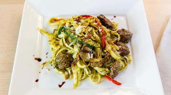Fettuccine pasta with pesto, red peppers, zucchini, mushrooms, beef, and parmesan cheese on a white plate.