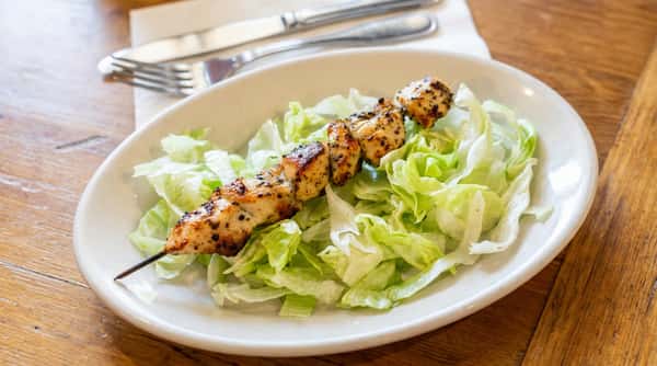 Chicken skewer with grilled, seasoned pieces of chicken and a side of shredded green lettuce on a white plate.