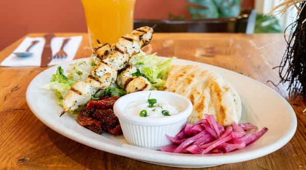 Chicken souvlaki plate with grilled chicken skewers, pita bread, Greek salad, tzatziki, sun-dried tomatoes, and pickled onions.