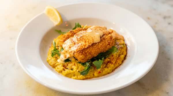 Fried fish with sauce and paprika on yellow grits with arugula, garnished with a lemon wedge.