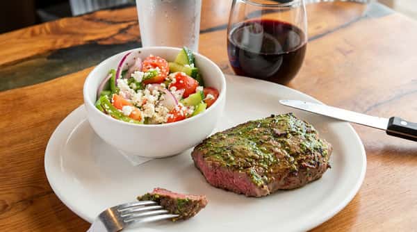 Medium-rare steak with green sauce, a Greek salad, and drinks on a wooden table.