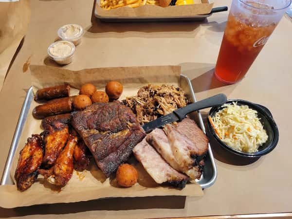 Loaded BBQ platter with smoked brisket, pulled pork, sausages, barbecue chicken wings, hush puppies, and creamy coleslaw, served with a drink — a true Southern barbecue feast.