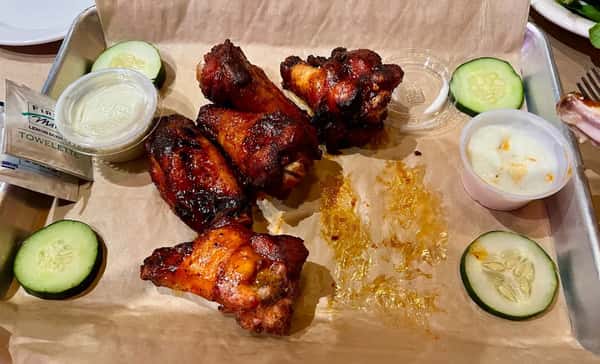 Char-grilled barbecue chicken wings with a caramelized smoky glaze, served with dipping sauces and fresh cucumber slices on a tray.