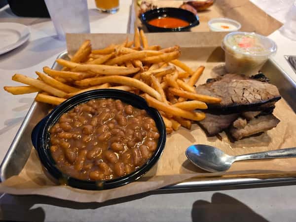 BBQ platter featuring tender smoked brisket slices, crispy golden French fries, sweet baked beans, and a side of creamy coleslaw — a classic American barbecue comfort meal served on a tray.