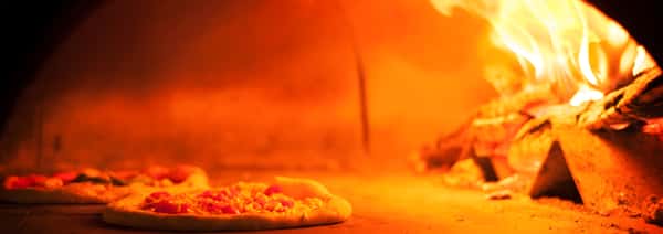A view inside a hot brick pizza oven, illuminated by bright orange flames in the background, with a pizza cooking on the stone surface.