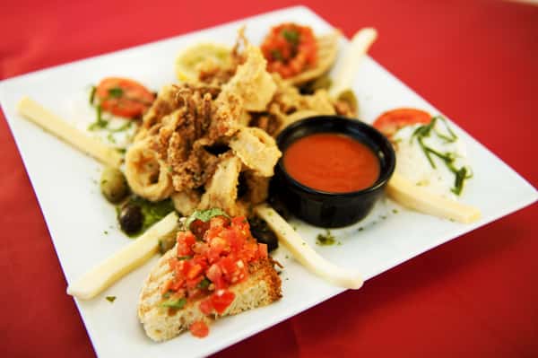 An overhead shot of a square white plate containing fried calamari, bruschetta, mozzarella sticks, and dipping sauce.