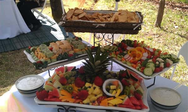 An outdoor shot of a buffet with a salad tray, a fruit tray, a cookies tray, and a wrap bites tray