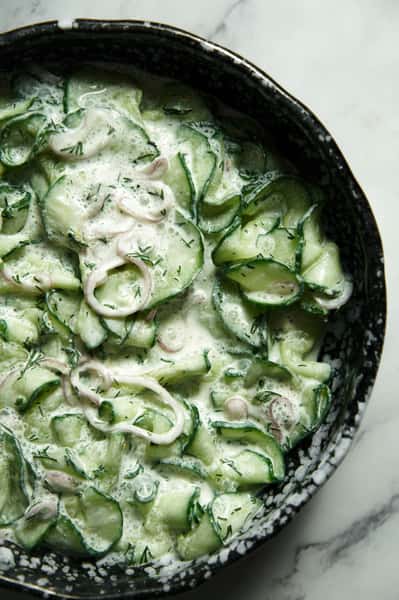 Creamy cucumber salad with thinly sliced cucumbers, shallots, and fresh dill in a tangy yogurt or sour cream dressing, served in a speckled black ceramic bowl.