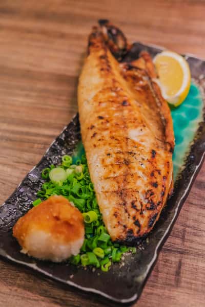 Grilled mackerel served on a black plate with chopped green onions, grated daikon, and a lemon wedge.