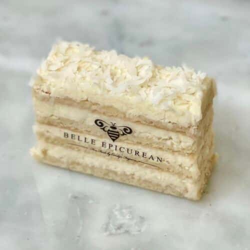 A rectangular slice of coconut layered cake with a branded logo on the side, topped with shredded coconut.