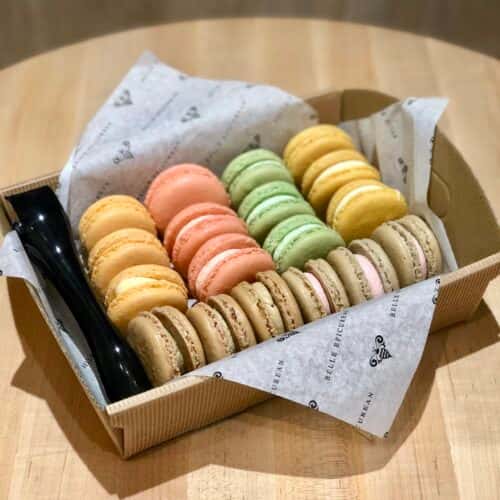 A box of colorful macarons in pastel shades of orange, green, pink, and yellow, arranged neatly with parchment paper lining.