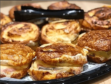 A tray of pastries, including sweet and savory croissant sandwiches with ham and cheese, alongside cinnamon rolls.