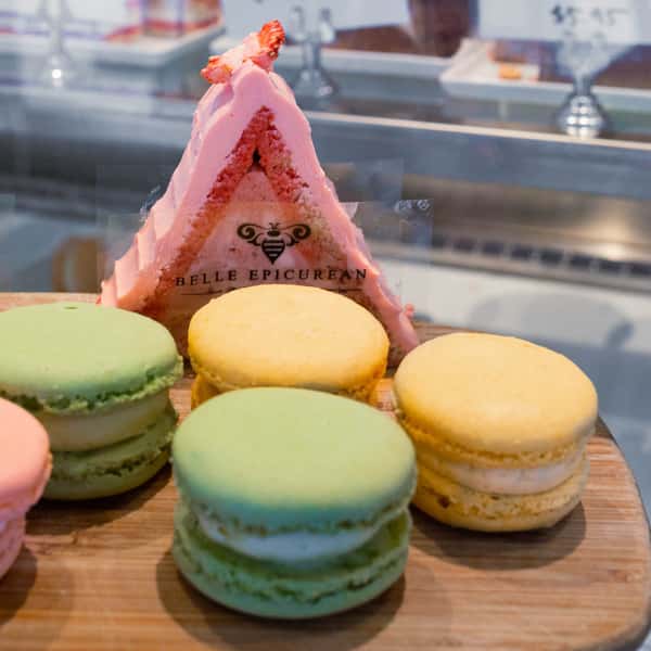 A display of colorful macarons in pastel pink, green, and yellow, with a triangular strawberry dessert featuring a branded logo in the background.